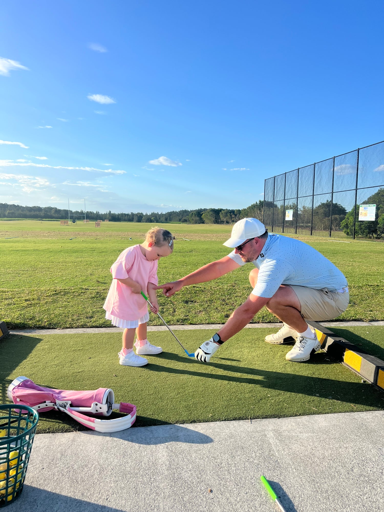 Dad teaching Daughter how to play golf at driving range. Pink toddler golf bag in the foreground.
